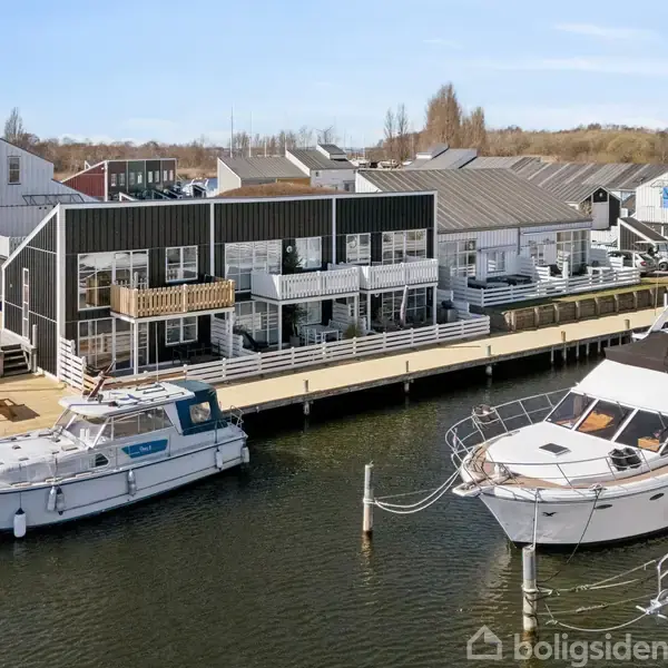 Både ligger ved kajen foran moderne huse med terrasser. Området har en blanding af marina og boliger, med åbne, grønne områder i baggrunden. Vandet er roligt og klart.