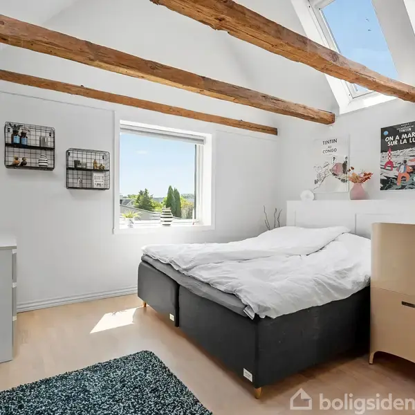 Soveværelse med sort seng placeret under to ovenlysvinduer. Træbjælker i loftet. Vindue med udsigt og plakater på væggen. Grønt tæppe på lyst trægulv. Planter på kommode.