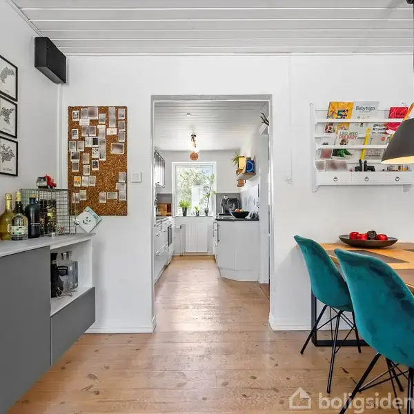 Spisebord med grønne stole står i en spisestue med trægulv. Væggene er dekoreret med billeder og en opslagstavle. Køkkenet ses gennem døråbningen i baggrunden.