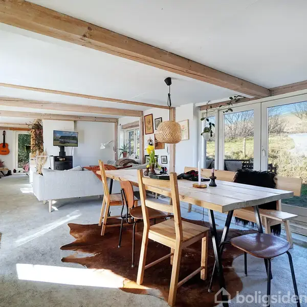 Spisebord står med stole omkring, belagt på et tæppe. Rummet har synlige træbjælker, en sofaområde i baggrunden og store vinduer med udsigt til en have. En guitar hænger på væggen.