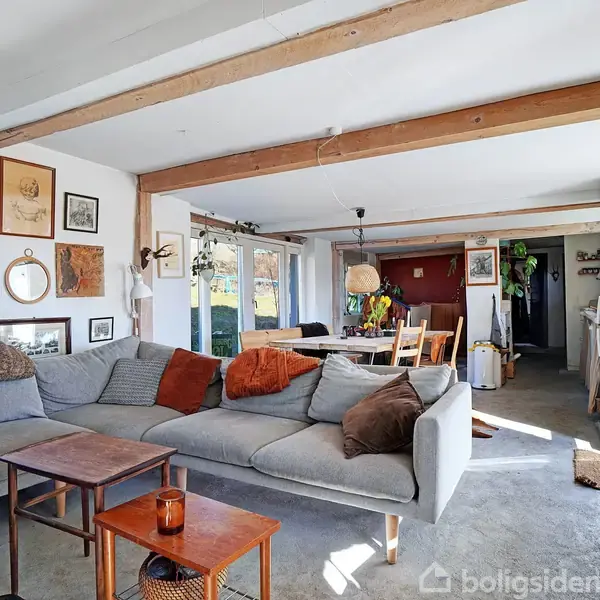 Stor grå sofa med puder står i en stue med træborde og vægkunst. Rundt om er en spiseplads med borde og stole, lyse vægge og store vinduer, der lader lys komme ind.