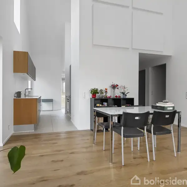 Et lyst køkken-alrum med spisebord og stole, trægulv og store vinduer til venstre, køkken med overskabe i baggrunden, omgivet af hvide vægge og grønne planter.