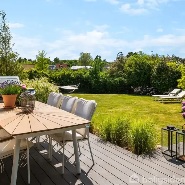Træbord med stole står på terrasse i en solrig have. Grill og blomster dekorerer området, mens en hængekøje og liggestole ses på plænen omgivet af buske og træer.
