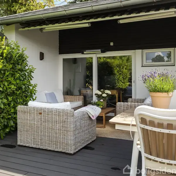 Terrassemøbler står på en træterrasse, hvor sofaer og et bord er arrangeret med puder og planter. Omgivet af grønne buske og en åben skydedør, der fører ind i huset.