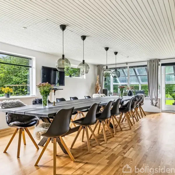 Langt spisebord med sorte stole omkring, dekoreret med planter, placerer i lyst rum med store vinduer der giver udsigt til grøn have. Hængelamper over bordet, trægulv og loftpaneler fuldender rummet.