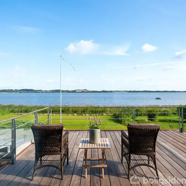 To kurvestole står på en træterrasse ud mod en stor sø eller fjord. Foran stolene står et lille bord med en plante. Græs og buske omgiver området.