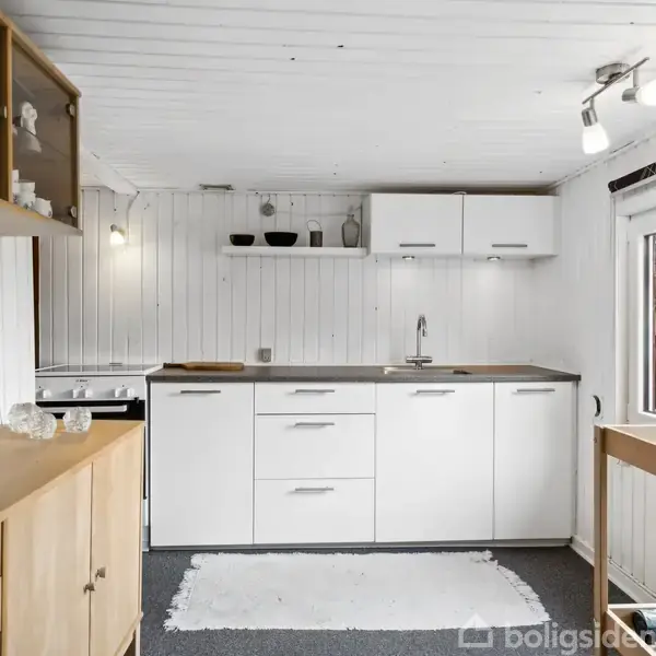 Hvidt køkken med lys træindretning, hvide skabe og hylder. En vask er centralt placeret. Et vindue giver udsigt til grønt udenfor. Teppens tekstur står i kontrast til gulvet.