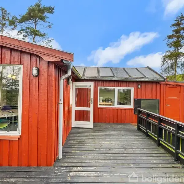 Rødt træhus med store vinduer står på en veranda med sort rækværk. Omkring huset er der træer og græsplæne under en blå himmel med spredte skyer.