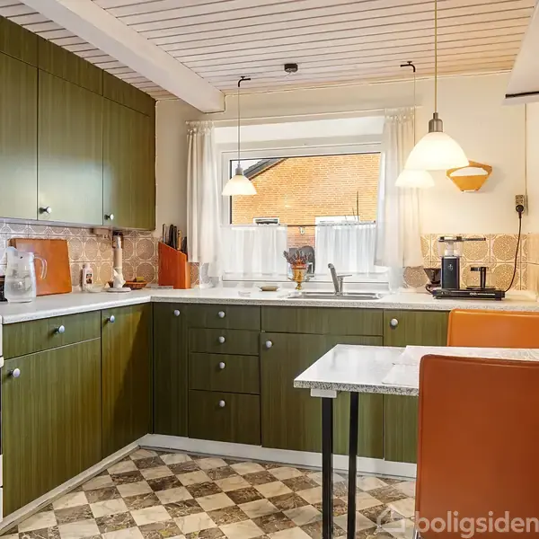 Køkken med grønne skabe og hvid komfur, kaffemaskine og køkkenudstyr. Lille bord med orange stole under hængelamper. Ternede gulvfliser og vindue med lysgardiner, der tilfører naturligt lys.