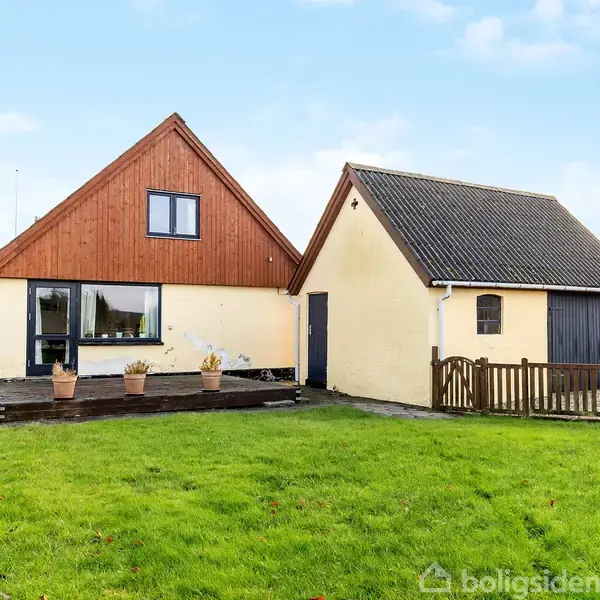 Hus med skråt tag står ved siden af en garage. Foran ses en græsplæne og terrasse med blomsterkrukker. Omgivet af andre bygninger i et beboelseskvarter.