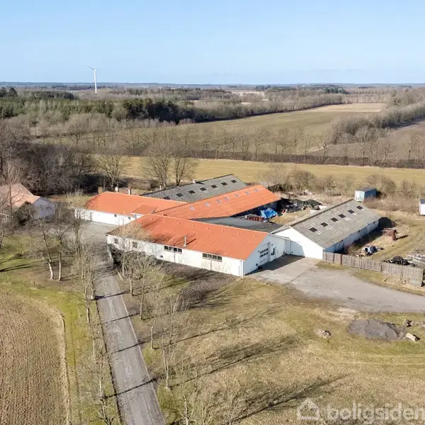 Stort landkompleks med flere langstrakte bygninger med rødt tag ligger på en åben mark omgivet af træer og marker. En vindmølle ses i baggrunden.