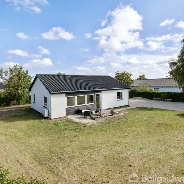 En hvid en-etagers bolig med sort tag står stille på en stor, grøn græsplæne. Omkring huset er der enkelte træer og buske under en blå himmel med skyer.