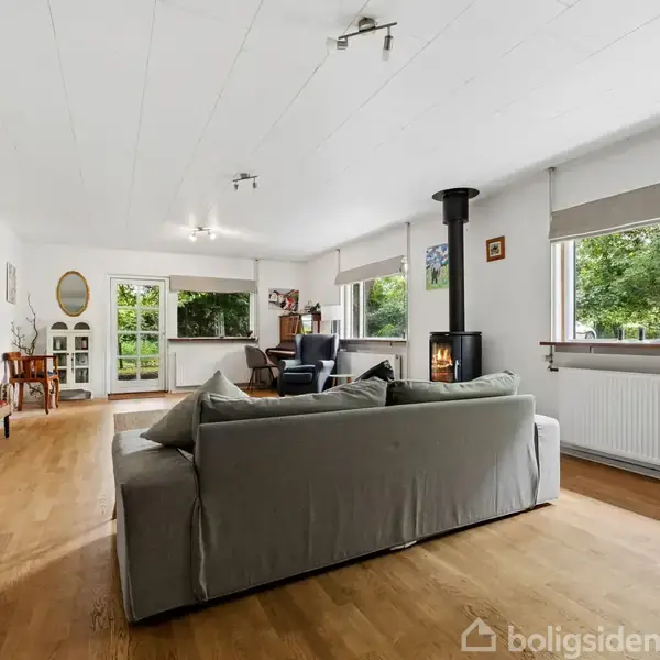 Grå sofa står i en lys stue med trægulv. Pejs brænder i hjørnet nær store vinduer, som giver udsigt til grøn natur. Møbler inkluderer kommode og stol.