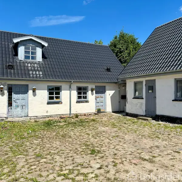 Et hvidt hus med sort tag ses fra gårdspladsen, omgivet af brolagte områder og græs. Flere døre og vinduer er synlige på facaden.