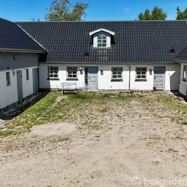 En gårdsplads omgivet af hvide bygninger med sorte tage i en hesteskoform. Midten er dækket af grus, mens der er lidt græs langs kanterne. En bænk står foran midterbygningen.