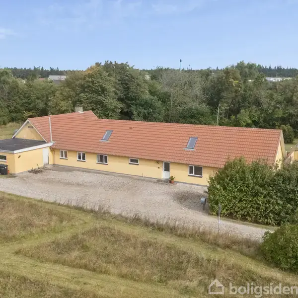 Et langt, gulmalet hus med rødt tag står i en naturlig omgivelser af træer og græs. En grusvej løber langs huset, og der er en lille tilbygning.