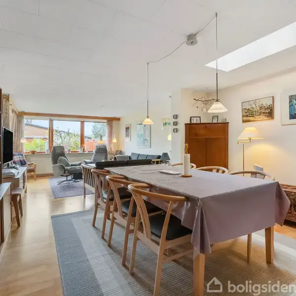 Spisebord med stole dækket af dug, opstillet i et lyst stueområde. Der er lamper hængende over bordet, kunstværker på væggen og store vinduer med udsigt.