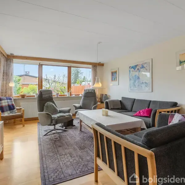 Stue med grå sofa og to lænestole omkring et hvidt sofabord. Store vinduer giver udsigt til have. Vægge dekoreret med kunst, og gulvet er i lyst træ.