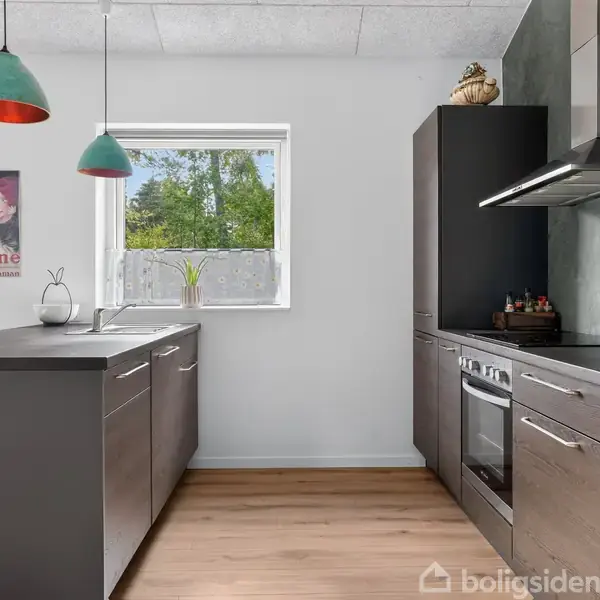 Køkken med mørke skabe og indbygget ovn, dekorativ plakat med teksten "Blédine En Semaine Maman" på væggen. Grønne pendellamper hænger over køkkenøen; lysindfald fra vindue.