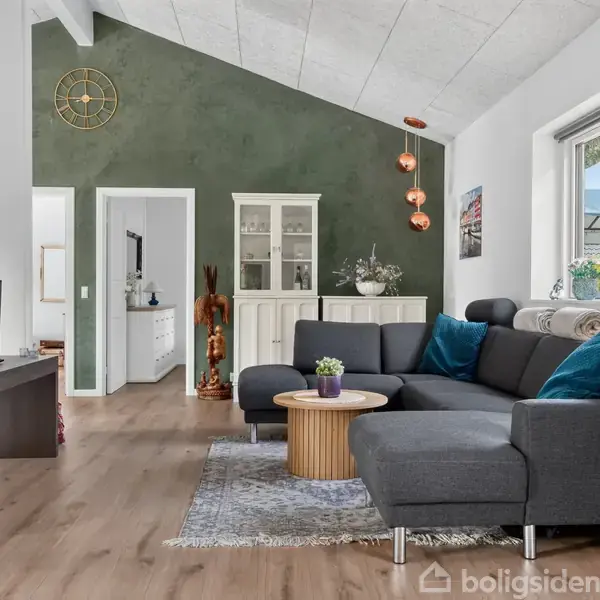 Grå sofa med farverige puder står på et tæppe i en stue. Et rundt bord er foran sofaen. En vægskænk og planter pynter rummet, der har en stor vindue med udsigt.