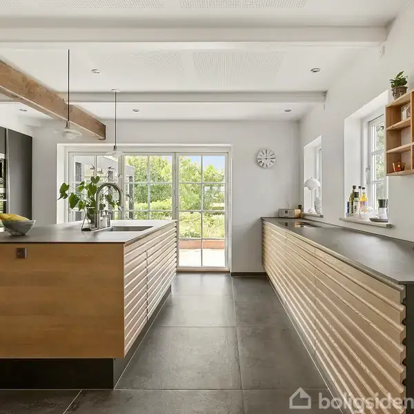 Køkken med en central ø i træ ser ud til at have moderne apparater, planter, og store glasdøre mod en have. Lyse vægge og mørke gulvfliser fuldender det stilrene design.