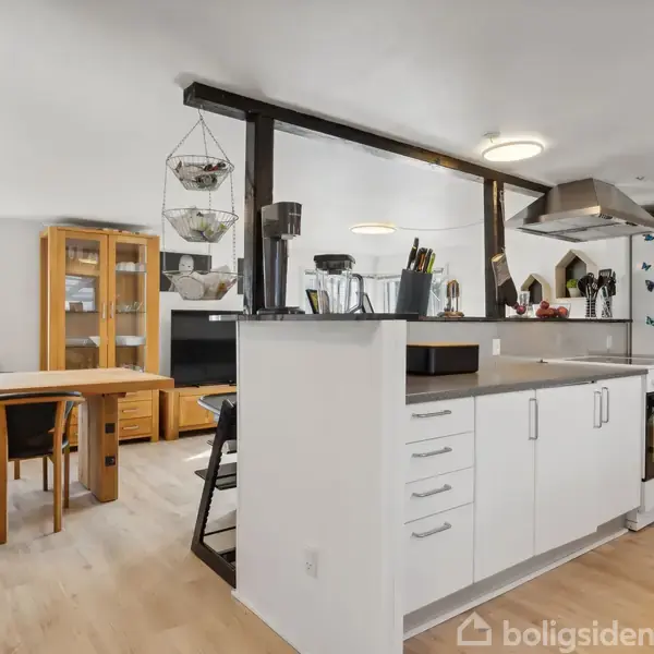 Køkken med hvide skabe og sort bordplade. Køleskab dekoreret med sommerfugle, ved siden af gasblus og emhætte. I baggrunden står et spisebord med træstole foran vindue.