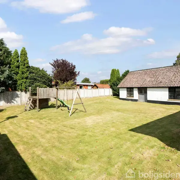 Legeplads med klatrestativ og rutsjebane står på græsplæne ved siden af et hvidt hus med gråt tag. Omgivet af hegn og træer under en blå himmel med skyer.