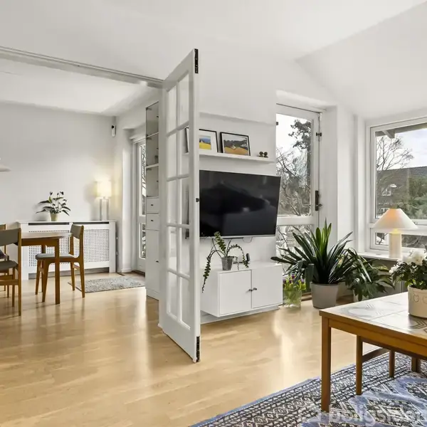 Stue med sofa og sofabord, placeret ved store vinduer med udsigt. Fjernsynet hænger på væggen, omgivet af planter. Åbne franske døre fører til spisestue med spisebord og stole.