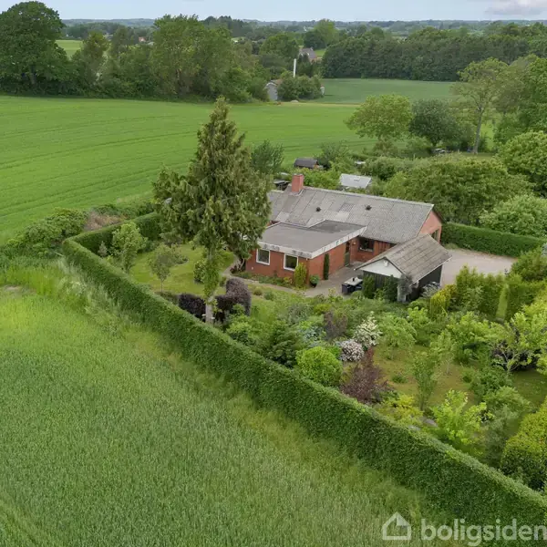 Et rødt murstenshus med gråt tag ligger omgivet af en frodig have med træer og buske. Det er placeret i et landbrugsområde med marker og grønne områder omkring.