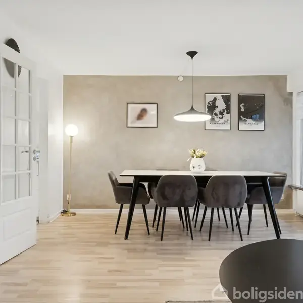 Et spisebord med sorte stole står i en lys spisestue. På væggen hænger kunstplakater, og en pendellampe hænger over bordet. Rummet har trælaminatgulv og et stort vindue.