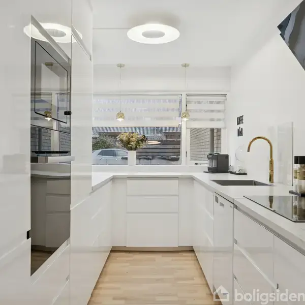 Et moderne køkken med hvide skabe og trælaminatgulv. Komfuret er indbygget i bordpladen, og der er en ovn til venstre. Store vinduer giver naturligt lysindfald.