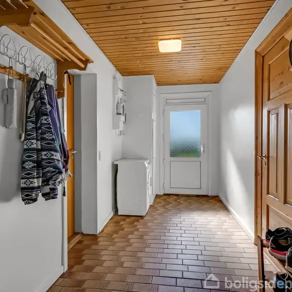 Gang med trægulv og træloft, jakker hængende på knagerække, sko opstillet ved en dør med mat glas. Vaskemaskine står i et hjørne.