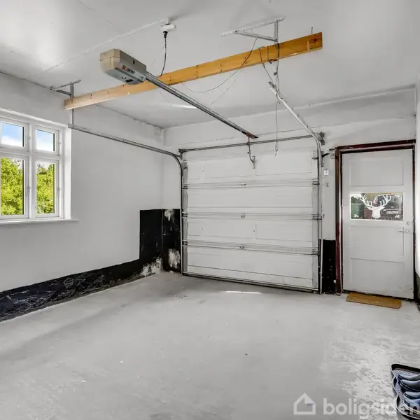 En garage med en garageport, der er lukket. Der er et vindue til venstre, som slipper naturligt lys ind. Vægge og gulv er hvide med sorte detaljer forneden. En radiator er monteret på højre væg.