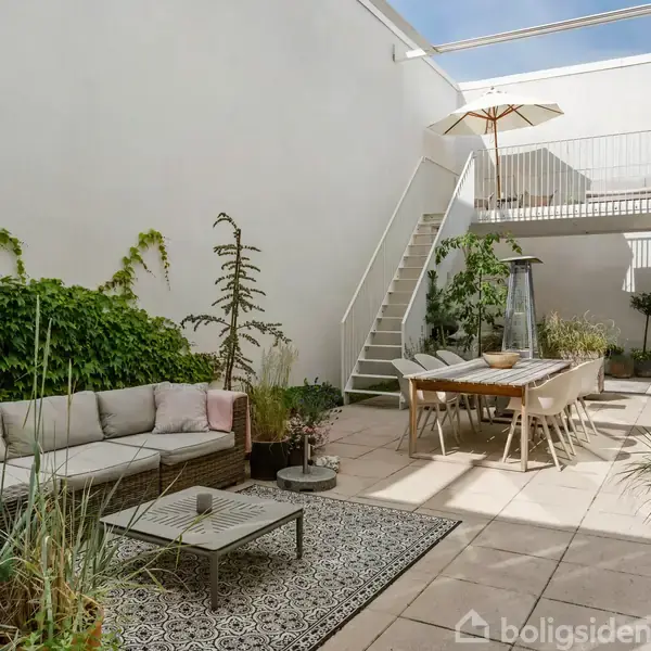 En gårdhave med sofa og bord på en tæppebelagt flisegulv. Ved siden af står et spisebord med stole. Muren er beklædt med klatreplanter, og en trappe fører til en balkon.