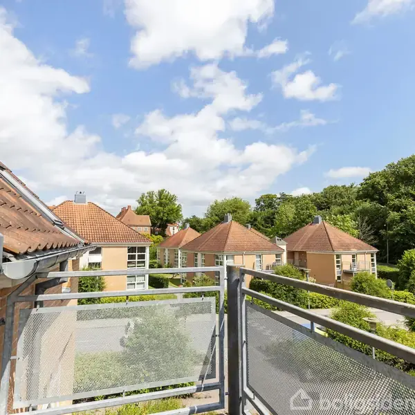 Altan med metalrækværk viser udsigt over tagbelagte rødstensbygninger og grønne træer under en blå himmel med spredte skyer.