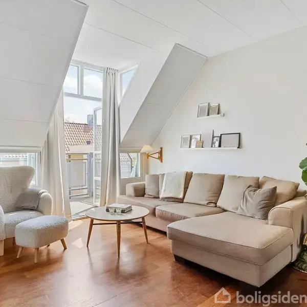 En beige hjørnesofa står i stuen ved siden af en stor plante og en Buddha-figur. En lænestol med skammel står foran vinduer med lyse gardiner, der slipper lys ind.