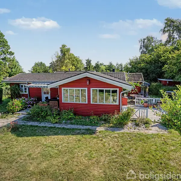 Rødt træhus står stille i en frodig have omgivet af store træer og buske en solskinsdag, med terrasse og grønne plæner omkring.