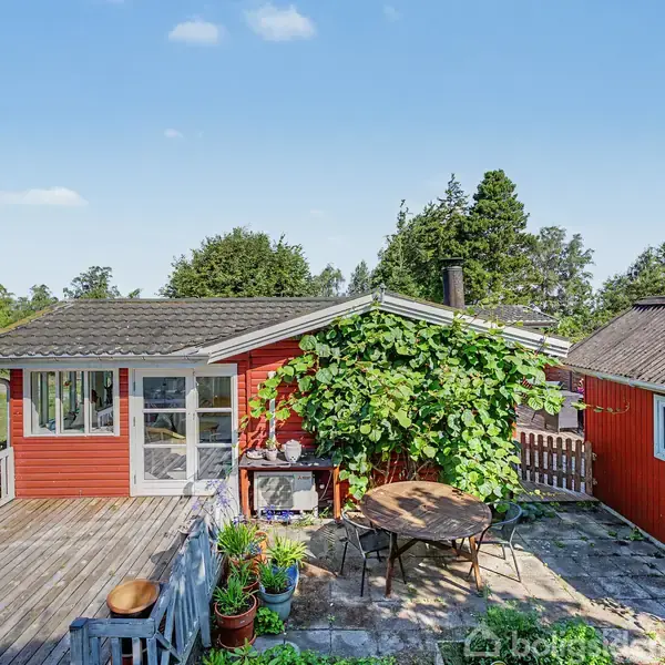 Rødt sommerhus med terrasse og klatreplanter, beliggende i en grøn have med træer og et gyngestativ. Border og stole står på terrassen, ideelt til udendørs ophold.