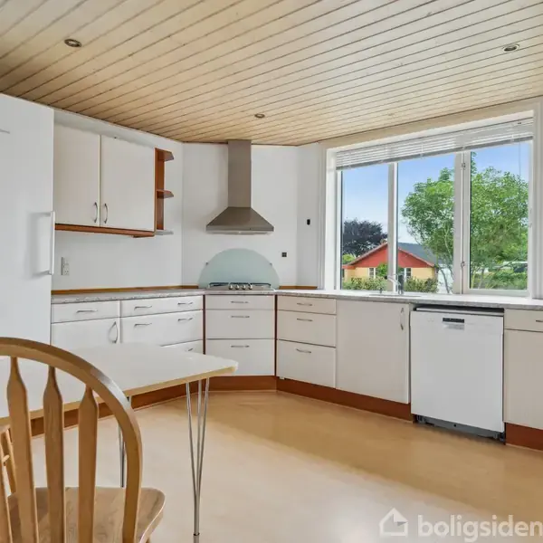 Køkken med hvide skabe og en skinnende opvaskemaskine under vinduet. Et køkkenbord med træstole står i forgrunden, mens store vinduer viser en have med træer udenfor.