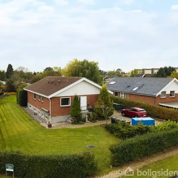 En rød murstensvilla med hvidt tag står på en velplejet græsplæne. En indkørsel med biler ses ved siden af, og huset er omgivet af hække i et villakvarter.