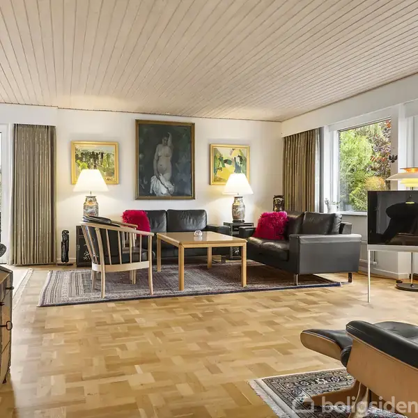 Stue med trægulv, hvor to sorte sofaer og et træbord danner siddeområde på et tæppe. Store vinduer viser træer udenfor. Flere lamper og malerier pryder væggene.