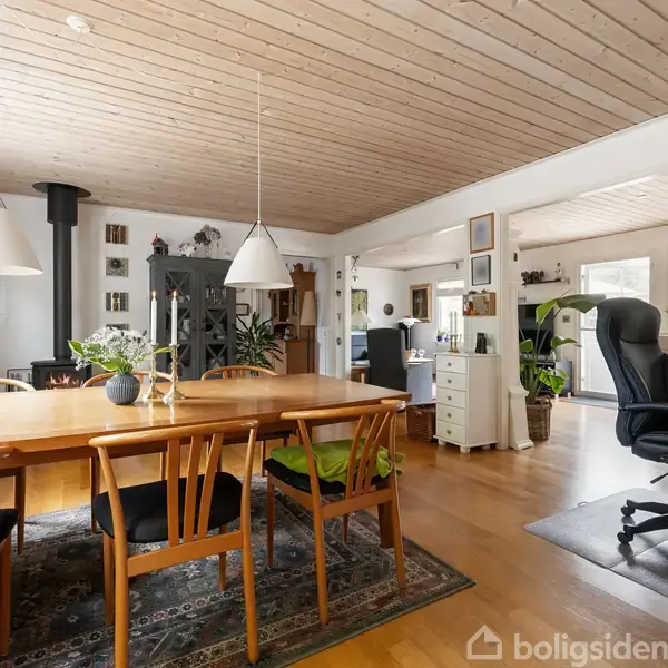 Et spisebord af træ er omgivet af stole og dekoreret med planter i potter; rummet har en brændeovn, skriveborde og flere grønne planter i en åben, lys stue.
