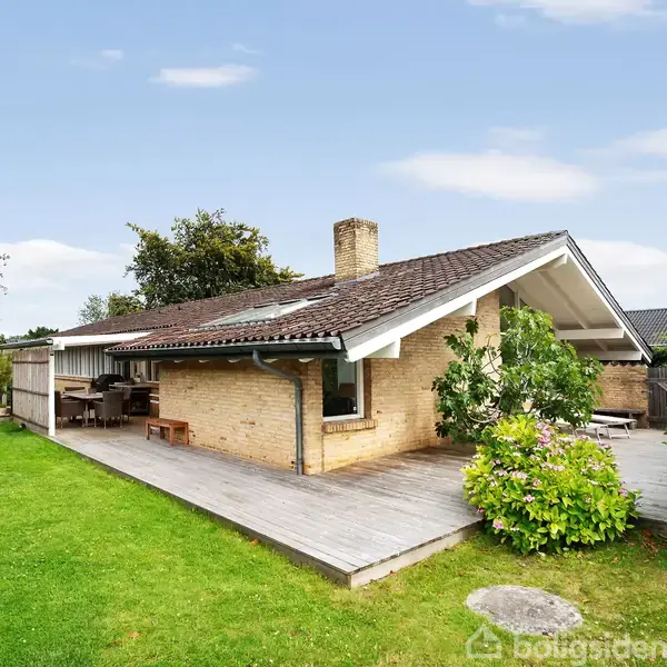 Et enfamilieshus i mursten står på en grøn græsplæne med en stor træterrasse. Terrassebord og bænke er til stede. Omkring huset er der buske og træer.