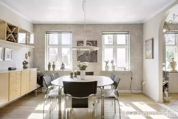 Et spisebord med sorte stole er placeret i et lyst rum med store vinduer, der lukker dagslys ind. Langs væggen står en skænk med dekorative genstande og hylder.