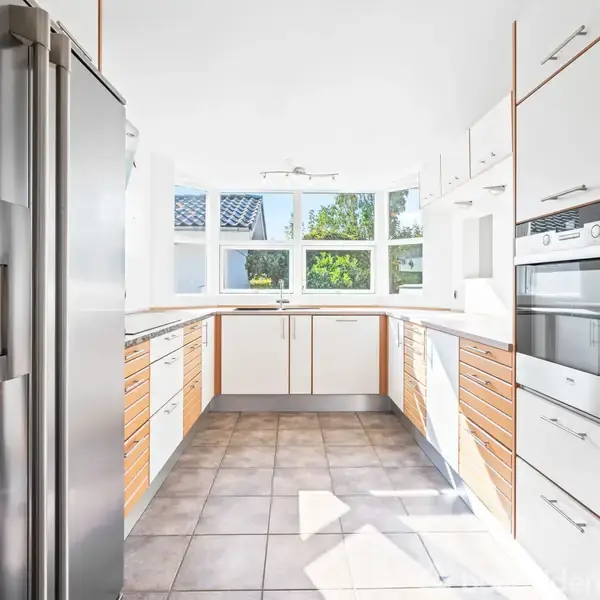 Køkken med stål køleskab og indbygget ovn, har hvide og træfarvede skabe og skuffer, kigger ud mod vinduer med udsigt til grønne træer, lyse fliser på gulvet.