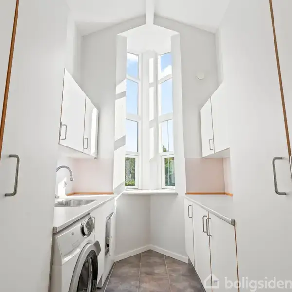 Vaskemaskine og tørretumbler står side om side i et smalt vaskerum med hvide skabe og grå gulvfliser. Store vinduer lukker lys ind fra den ene ende.