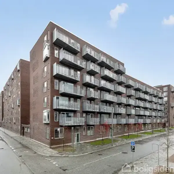 Bygning med flere etager og altaner på alle niveauer, står langs en bred vej i et boligkvarter. Der er cykelstativer og parkeringspladser i forgrunden.