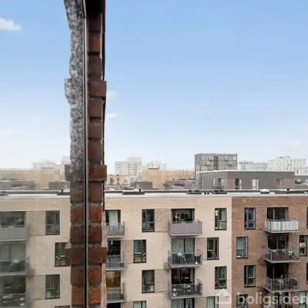 På billedet ses en moderne boligblok med altaner, set fra en høj bygning. I baggrunden er flere lignende bygninger under en klar blå himmel.