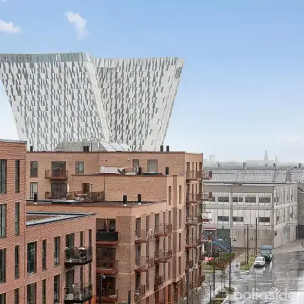 Etagedelejligheder med røde mursten står i forgrunden, mens en markant, moderne bygning med vinklet facade dominerer baggrunden under en blå himmel. En gade med biler ses mellem bygningerne.
