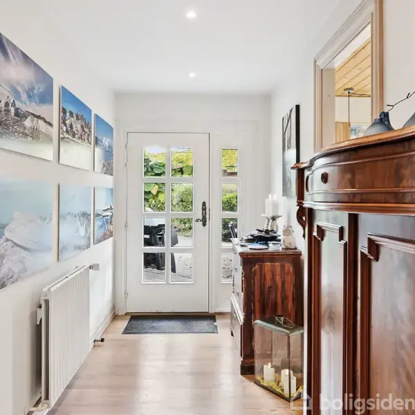 En lys gang med fotografier på venstre væg fører til en glasdør. En stor, antik trækommode står til højre. Der er fotografier af vinterlandskaber på væggen.